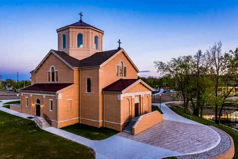 Twilight exterior of the brick oratory with its octagonal lantern tower lit from within, seen from above across the paved forecourt and campus lawns.