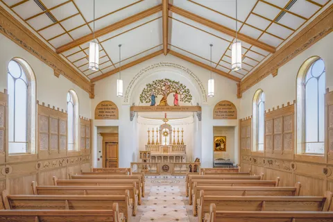 Interior view of a chapel nave looking toward a marble altar beneath a carved tree-of-life relief, flanked by wood-paneled walls and arched windows.