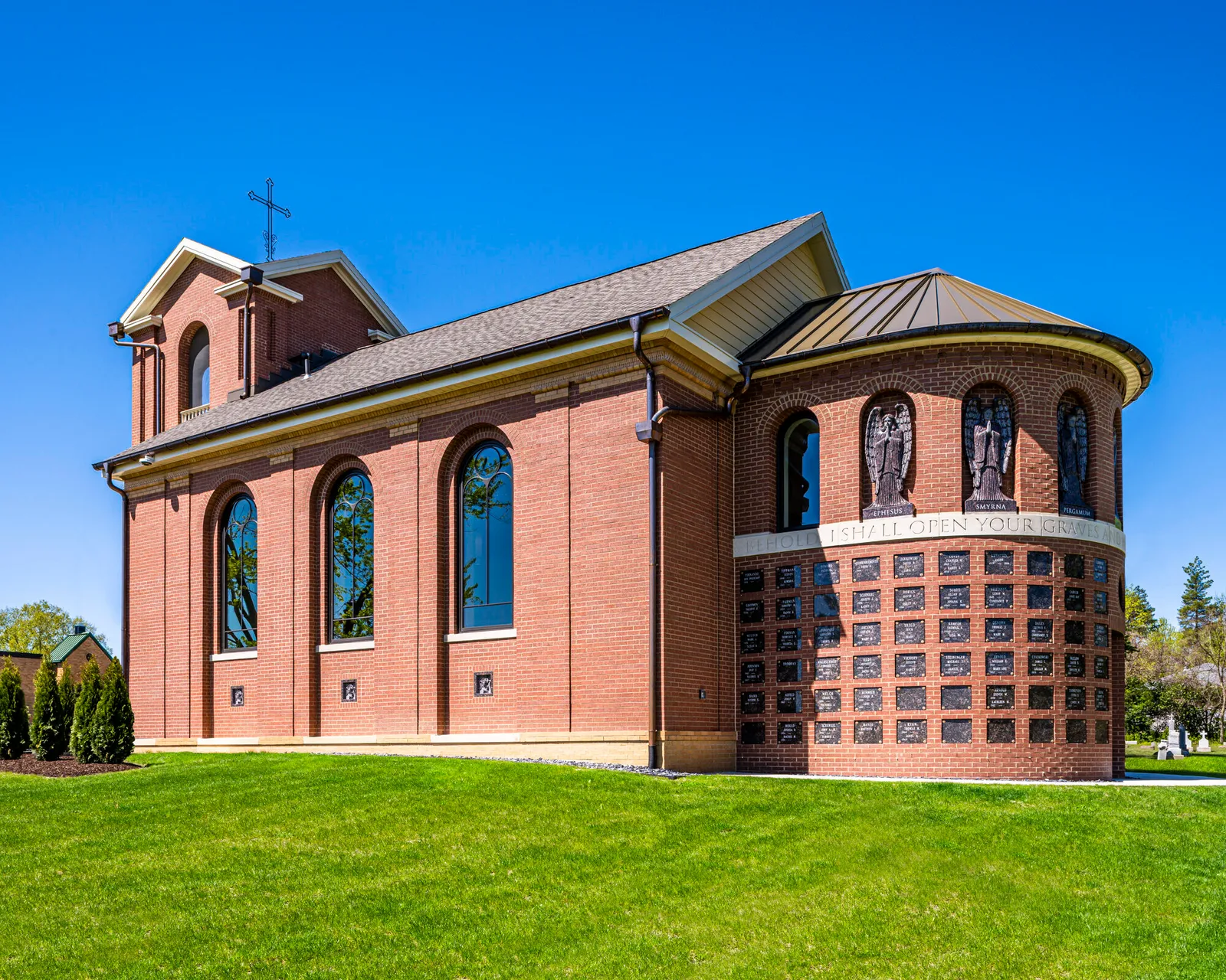 Exterior corner view of a brick chapel showing a curved columbarium wall with carved angel reliefs and inscribed memorial plaques under a blue sky.