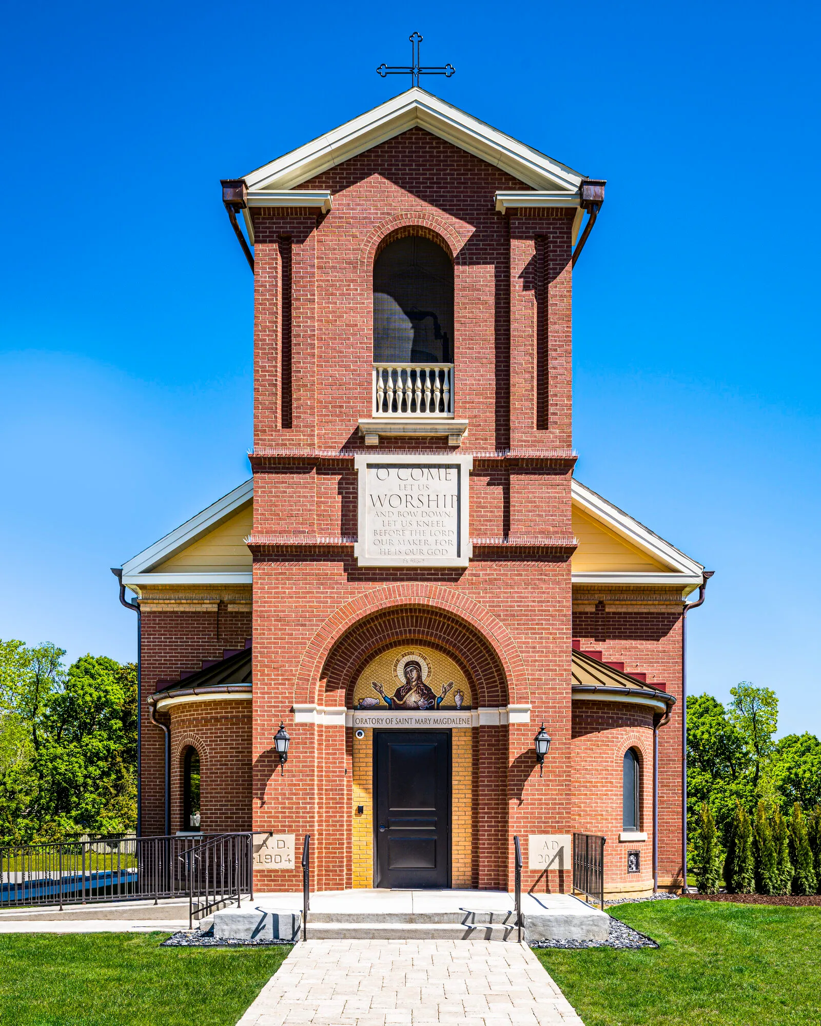 Symmetrical frontal view of a red brick chapel bell tower with a mosaic icon over the entrance door and an inscribed stone tablet above.