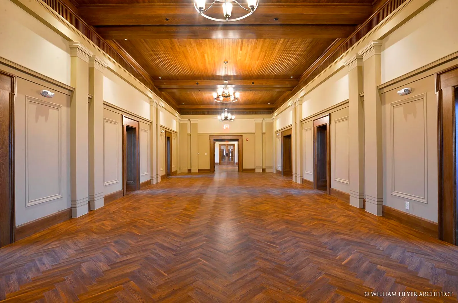 The completed John Paul II Center corridor: a herringbone wood floor between panelled walls with fluted pilasters, a coffered wood ceiling and brass chandeliers, doors to the faculty offices either side.