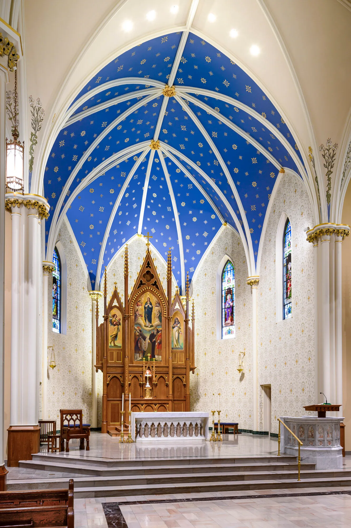 The sanctuary with a carved wood altarpiece depicting the Assumption, set beneath a blue star-patterned vault.